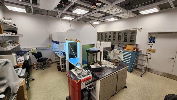 A table with various small containers sits in front of large contained workspaces that have overhead blueish light.